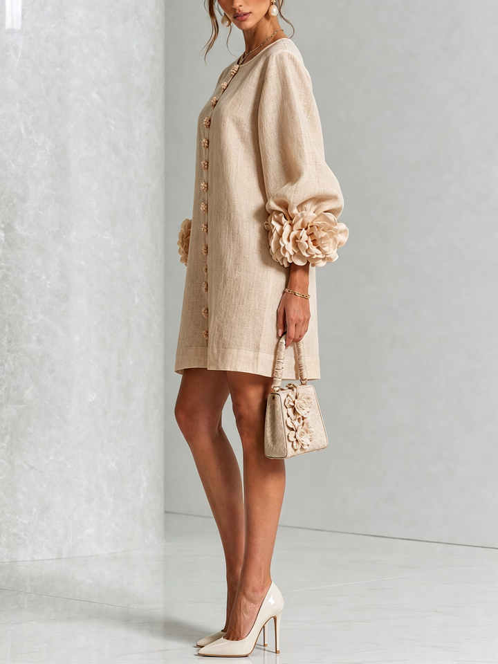 Model i beige minikjole set fra siden, med pufærmer, rosmanchetter og række af blomsterknapper, holder lille taske med blomsterpynt. 24