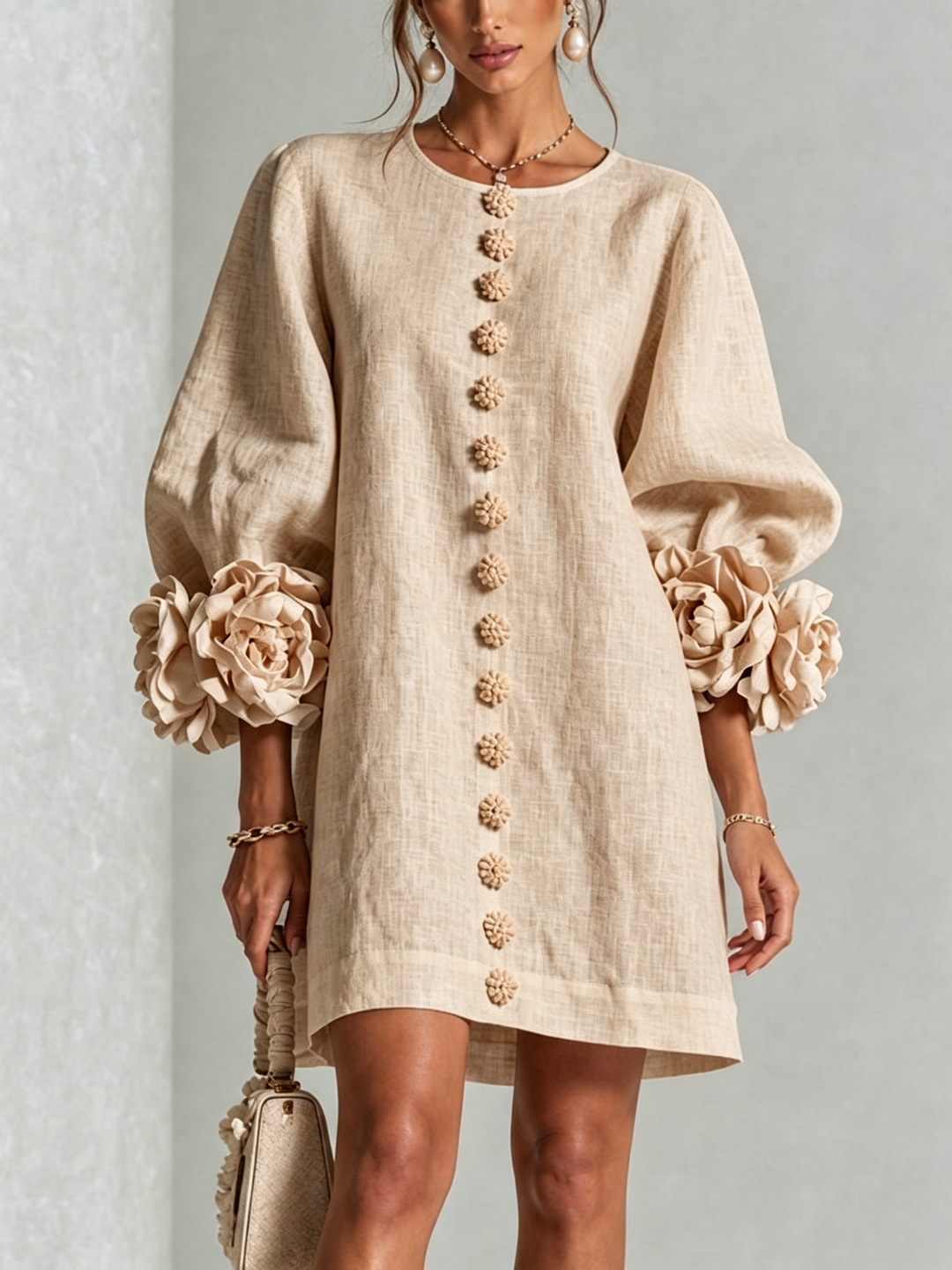 Nærbillede forfra af beige minikjole med række af blomsterknapper, pufærmer og store rosmanchetter, model med perleøreringe og lille taske. 23