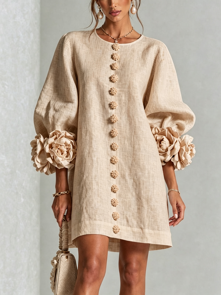 Nærbillede forfra af beige minikjole med række af blomsterknapper, pufærmer og store rosmanchetter, model med perleøreringe og lille taske. 23