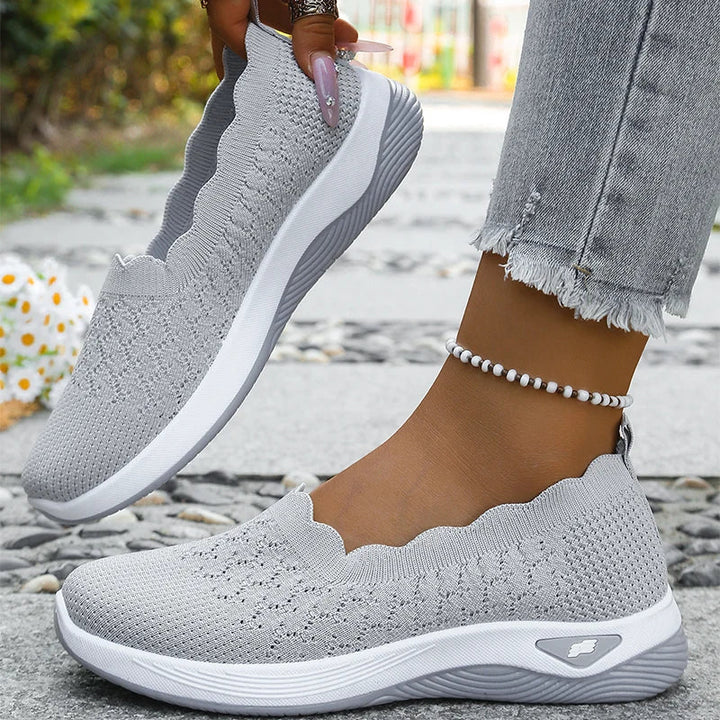 Grå strikkede slip-on-sko, den ene løftet i hånden og den anden på foden, med hvid sål 10