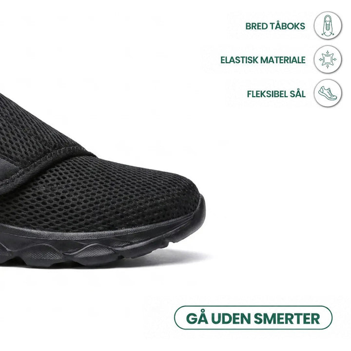 Nærbillede af den brede tåkappe på en sort mesh-sko med ikoner for bred tå, elastisk materiale og fleksibel sål. 14