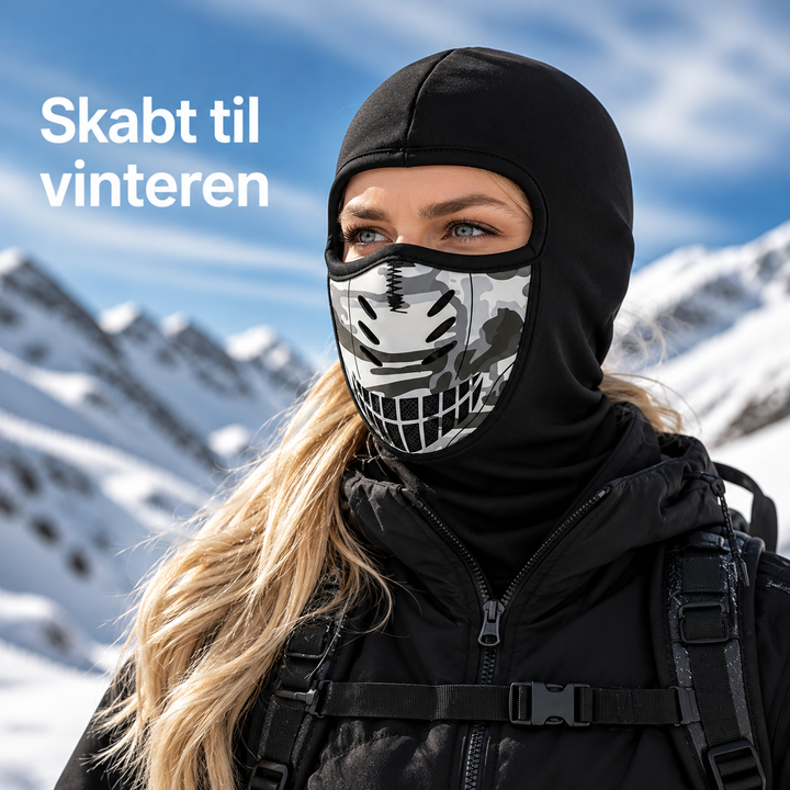 Kvinde med sort ansigtsmaske med camouflagepanel og ventilationshuller foran et snedækket bjerglandskab under blå himmel. 1