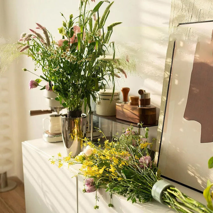 Vilde blomster i en metalvase på et hvidt skab, mens en anden buket med gule blomster ligger ved siden af med en grøn indsats. 3