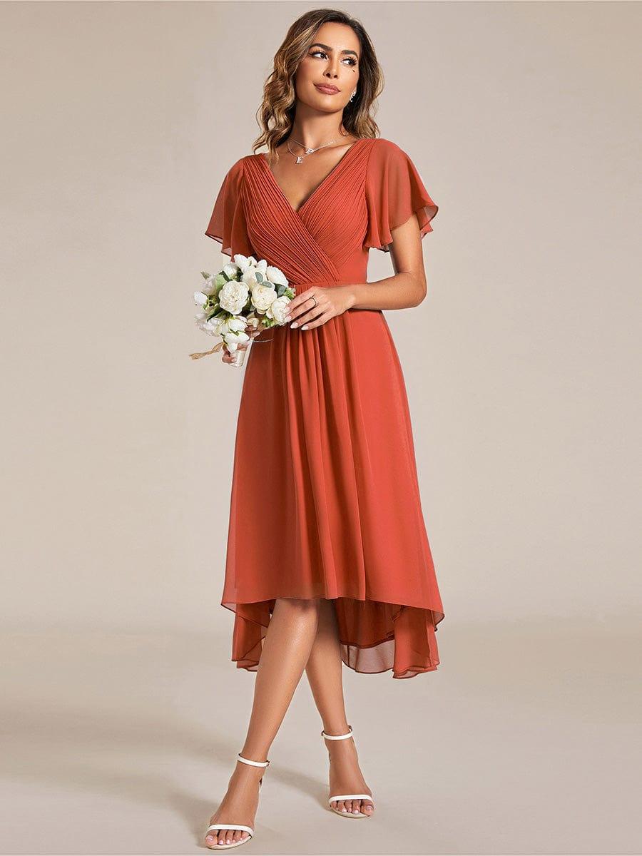 Model i rustorange chiffonkjole holder en lille buket hvide blomster, set skråt forfra med hvide sandaler. 95