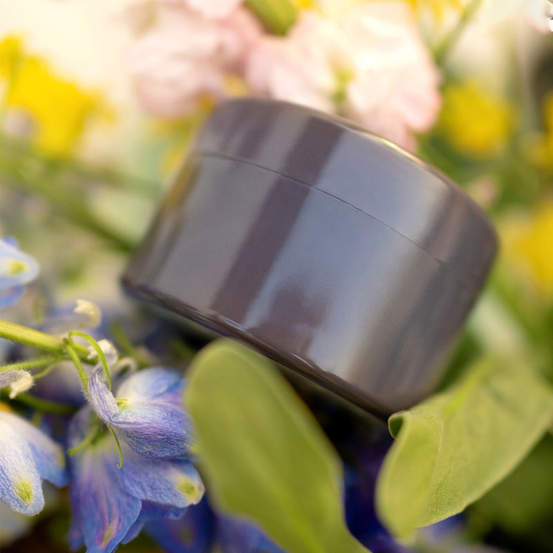 Mørkegrå, blank rund indsats set fra siden mellem blå blomster, grønne blade og sarte lyserøde og gule blomster. 16
