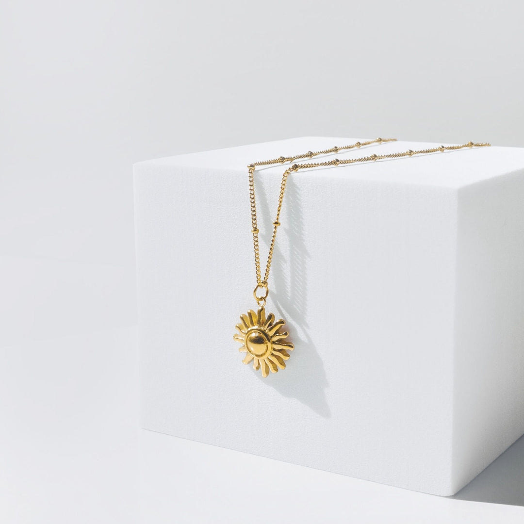 Goldene Sonnen-Halskette auf weißem Würfel, elegantes Schmuckdesign, modisches Accessoire.