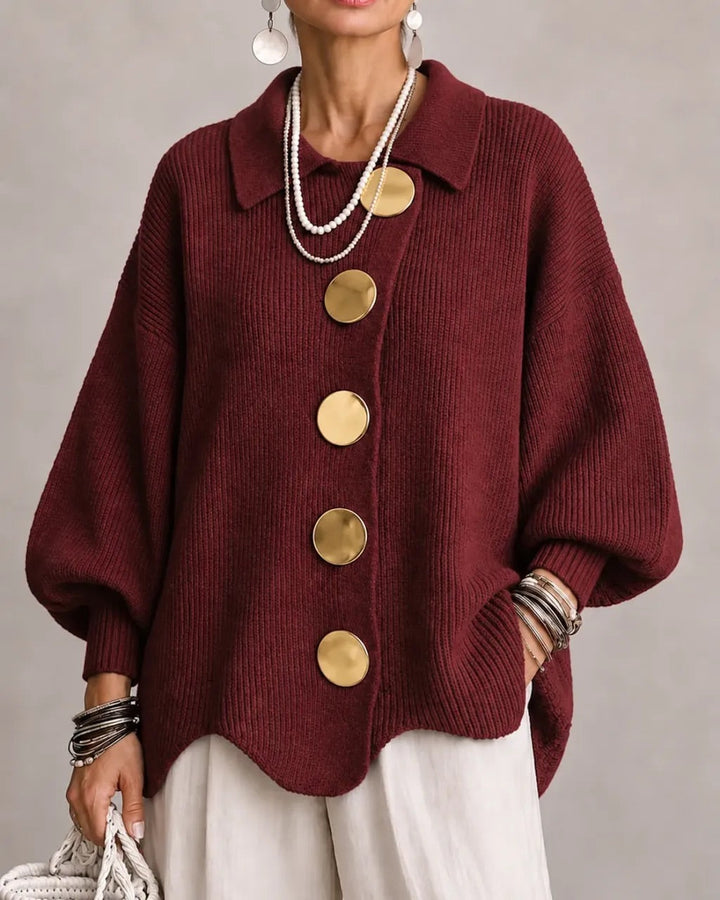 Model i bordeauxrød ribstrikket cardigan med store guldfarvede knapper, spids krave og bølgekant, båret med hvide bukser. 19