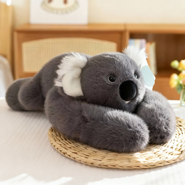 Grå koala-krammedyr med hvide ører og stor sort næse ligger med lange arme på et flettet underlag. 16