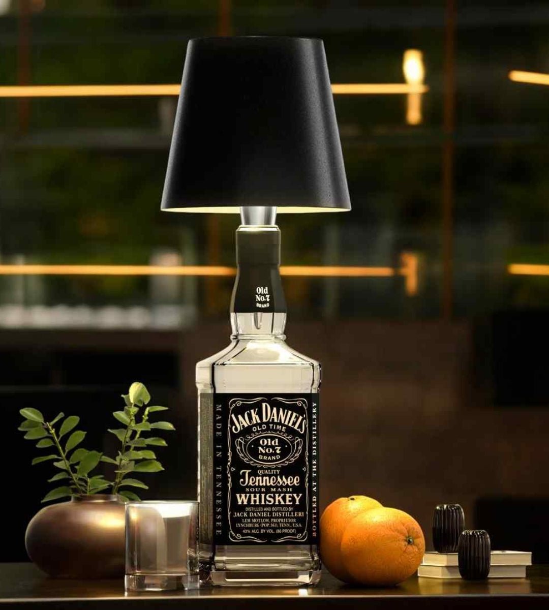 Sort lampeskærm på en kantet whiskeyflaske, stillet på et bord med glas, appelsiner og en plante i baggrunden. 2