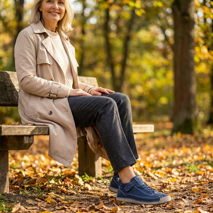 Smilende kvinde i beige trenchcoat sidder på en bænk i efterårsskoven og bærer mørkeblå gåsko med velcrolukning. 21