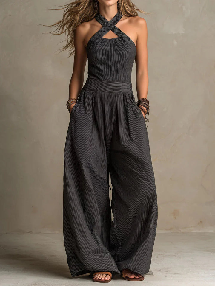 Model i mørkegrå, fintstribet jumpsuit med krydsede halsstropper, hænder i lommerne og meget vide ben, set forfra. 1