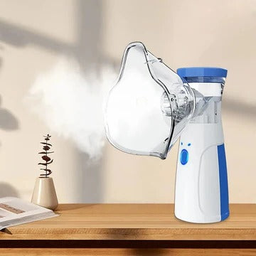 Hvid og blå håndholdt nebulisator med påsat maske, der udsender tåge, på et træbord ved siden af en bog og en lille plante. 1