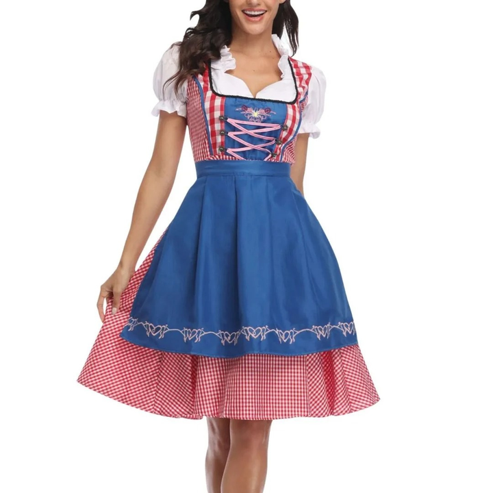 Model i dirndl med klarblåt forklæde over rød-hvidt ternet skørt, pink snøring i livet og hvide pufærmer, set forfra. 3