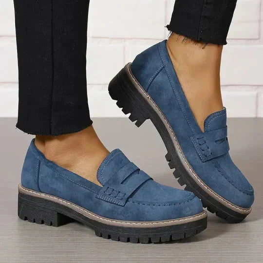Blå ruskindslignende loafers med tyk sort sål og beige kant, båret med sorte bukser med flossede ben. 9