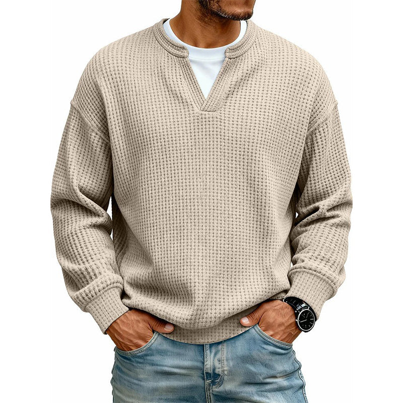 Mand i beige vaffelstrikket top med V-udskæring over hvid T-shirt, hænder i lommerne på jeans. 11