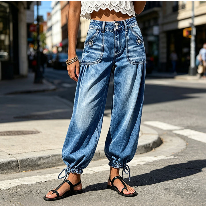 Model i blå tøndejeans, hvid blondetop og sorte sandaler på en solbeskinnet bygade, set forfra med bindebånd ved anklerne. 2