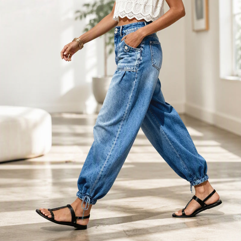 Model går i lyst rum i blå tøndejeans set fra siden med hånden i lommen og bindebånd ved sømmen. 7