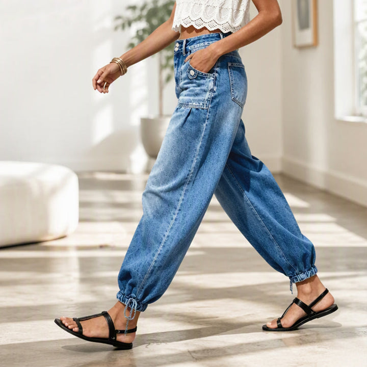 Model går i lyst rum i blå tøndejeans set fra siden med hånden i lommen og bindebånd ved sømmen. 7