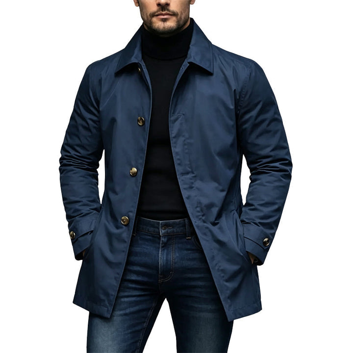 Mand i marineblå mellemlang trenchcoat med guldfarvede knapper, båret over sort rullekravetrøje og mørke jeans, med hænderne i lommerne. 4