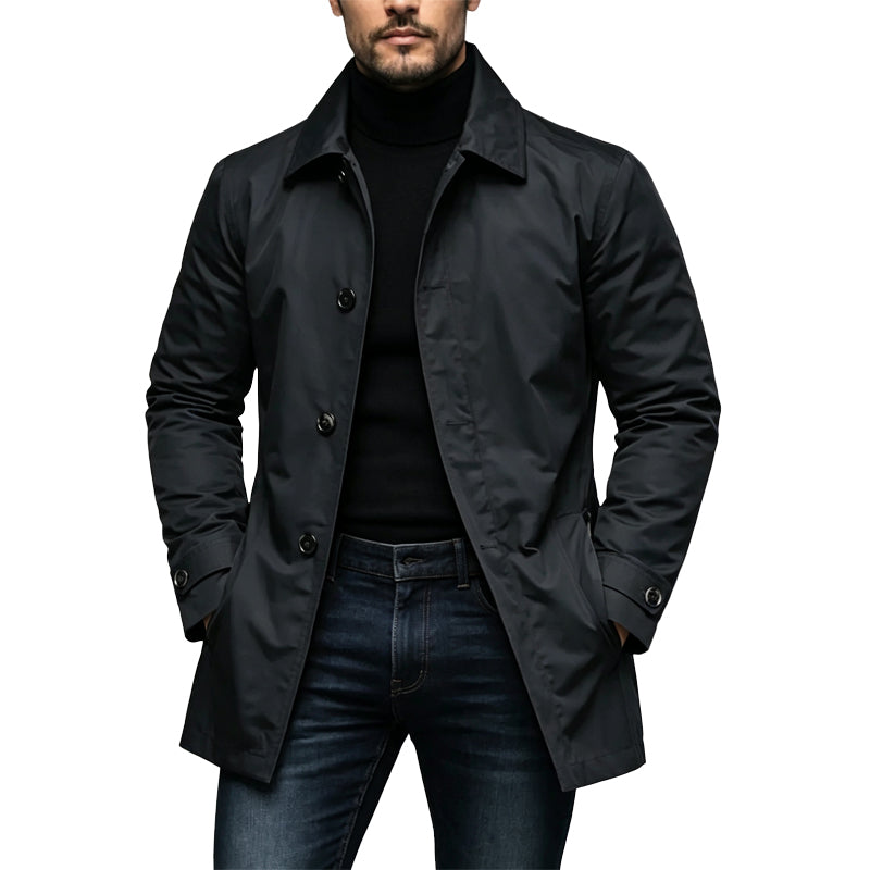 Mand i sort mellemlang trenchcoat med knapper og krave, båret over sort rullekravetrøje og mørke jeans, med hænderne i lommerne. 1