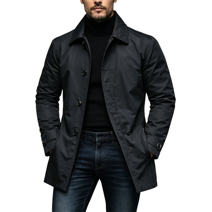 Mand i sort mellemlang trenchcoat med knapper og krave, båret over sort rullekravetrøje og mørke jeans, med hænderne i lommerne. 1
