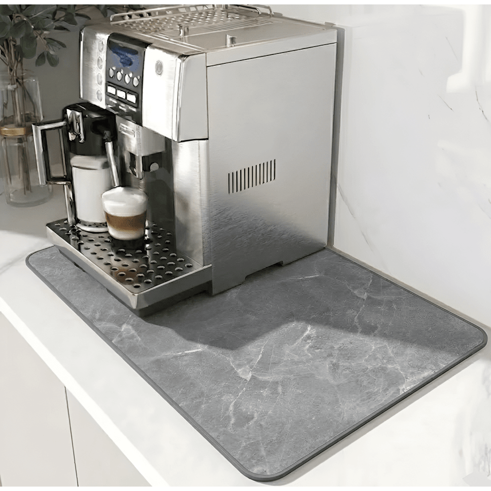 Grå måtte med marmormønster og mørkere kant under en kaffemaskine på en hvid bordplade. 12