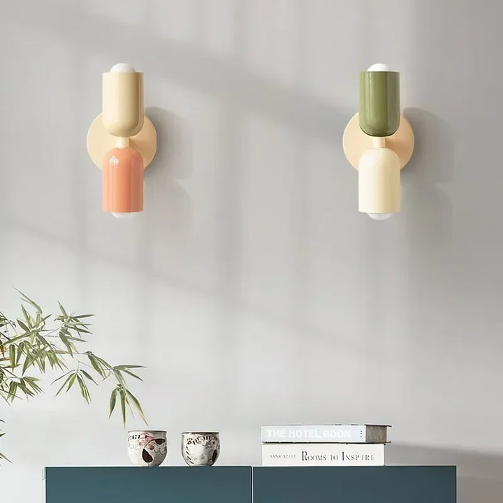 To væglamper i creme og fersken samt oliven og creme på grå væg over et grønt skænk med bøger, skåle og plantegrene. 14