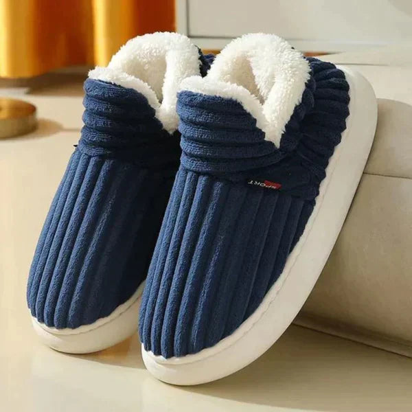Par riflede slippers i marineblå med hvid plysforing og tyk hvid sål, stillet på et lyst gulv ved en sofa. 1