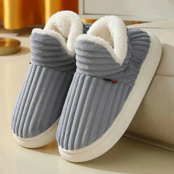 Par riflede slippers i grå med hvid plysforing og tyk hvid sål, stillet på et lyst gulv ved en sofa. 2