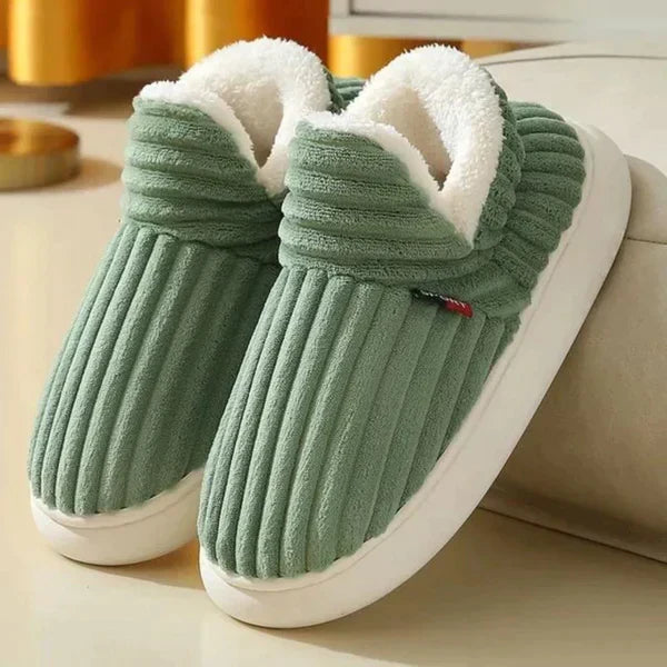 Par riflede slippers i grøn med hvid plysforing og tyk hvid sål, stillet på et lyst gulv ved en sofa. 4