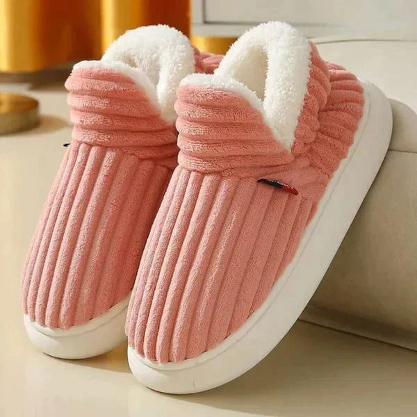 Par riflede slippers i laksrosa med hvid plysforing og tyk hvid sål, stillet på et lyst gulv ved en sofa. 5
