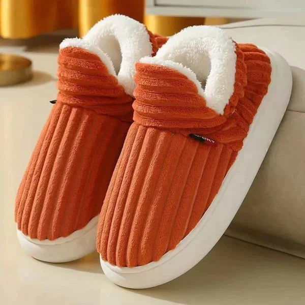 Par riflede slippers i orangerød med hvid plysforing og tyk hvid sål, stillet på et lyst gulv ved en sofa. 6