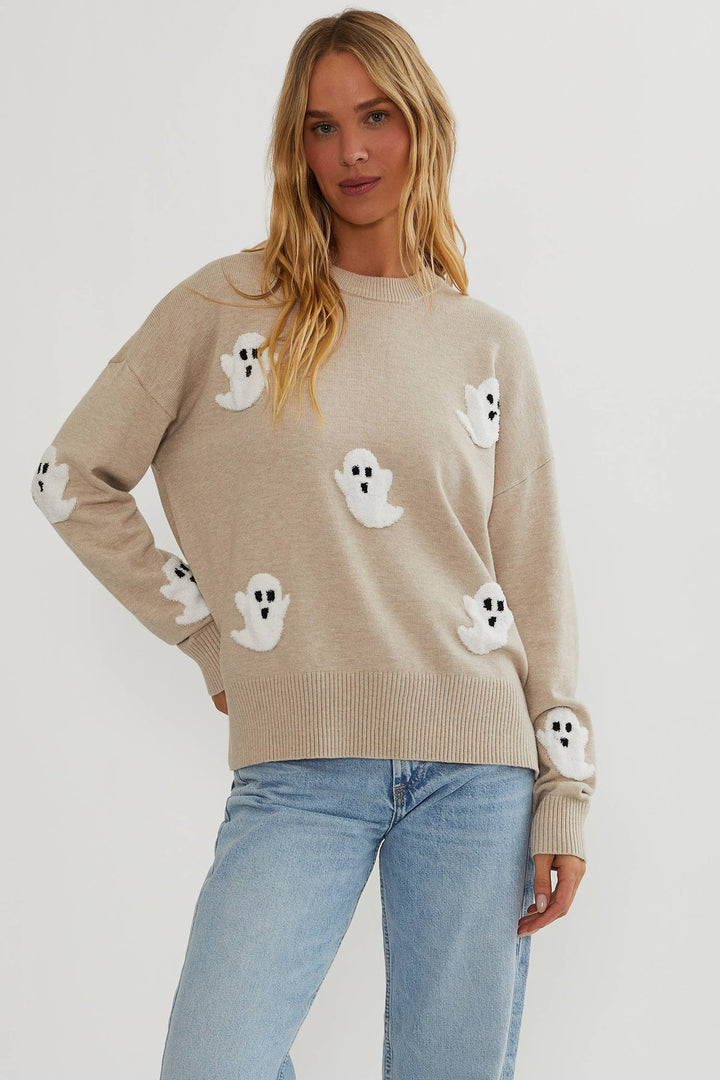 Model set forfra i beige strikket sweater med hvide, bløde spøgelsesapplikationer og ribkanter, båret til lyse jeans. 1