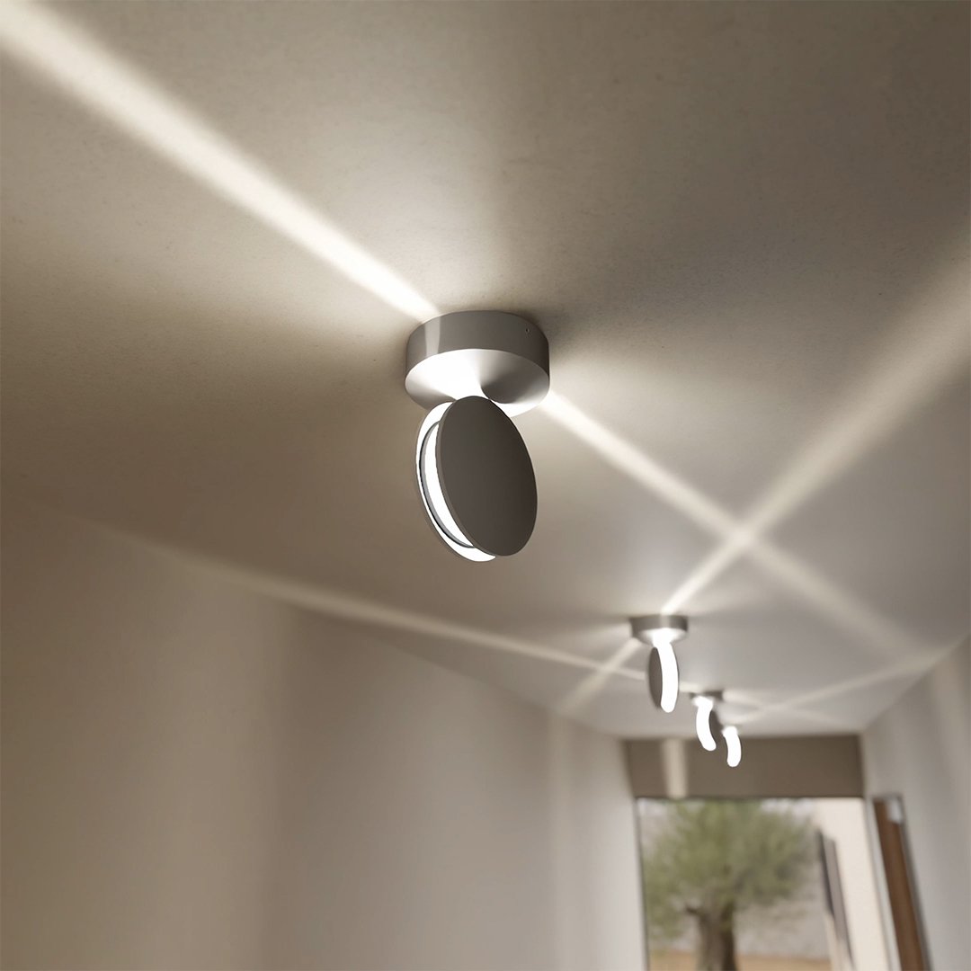 Hvid loftlampe med drejet hoved set fra siden, med krydsende lysstråler over loftet i en gang og flere lamper i baggrunden. 4