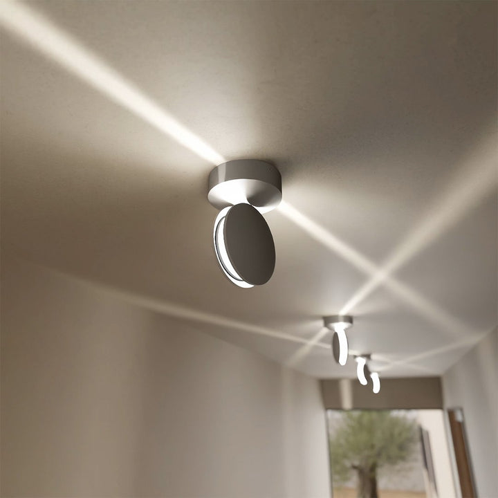 Hvid loftlampe med drejet hoved set fra siden, med krydsende lysstråler over loftet i en gang og flere lamper i baggrunden. 4